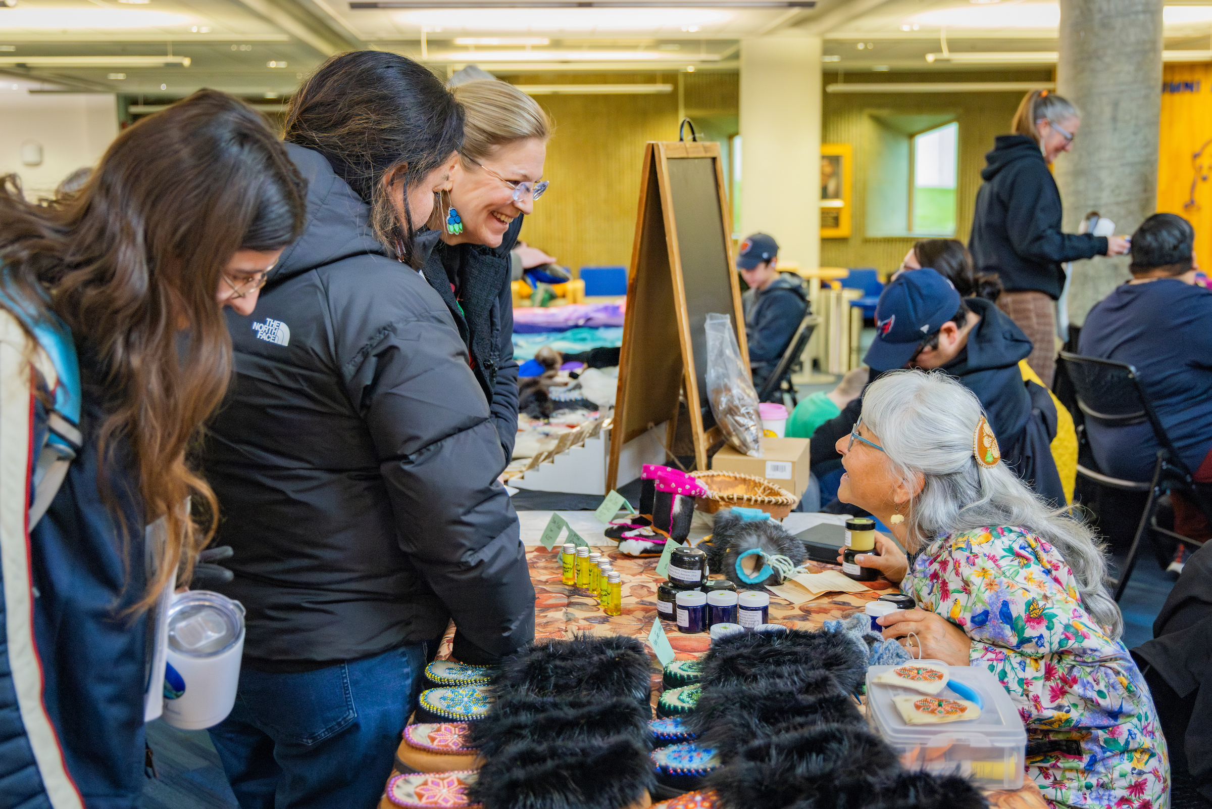 An Elder in a kuspuk sits at a table displaying beaded slippers and other goods, talking with three smiling people.