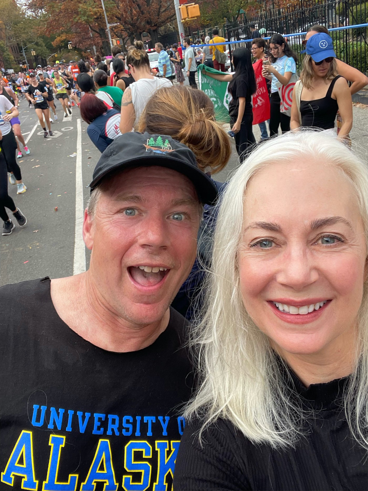 A man in a ball cap and a University of Alaska Fairbanks T-shirt, his face flushed from running, poses with a white-haired woman as marathon runners and spectators fill the background street.