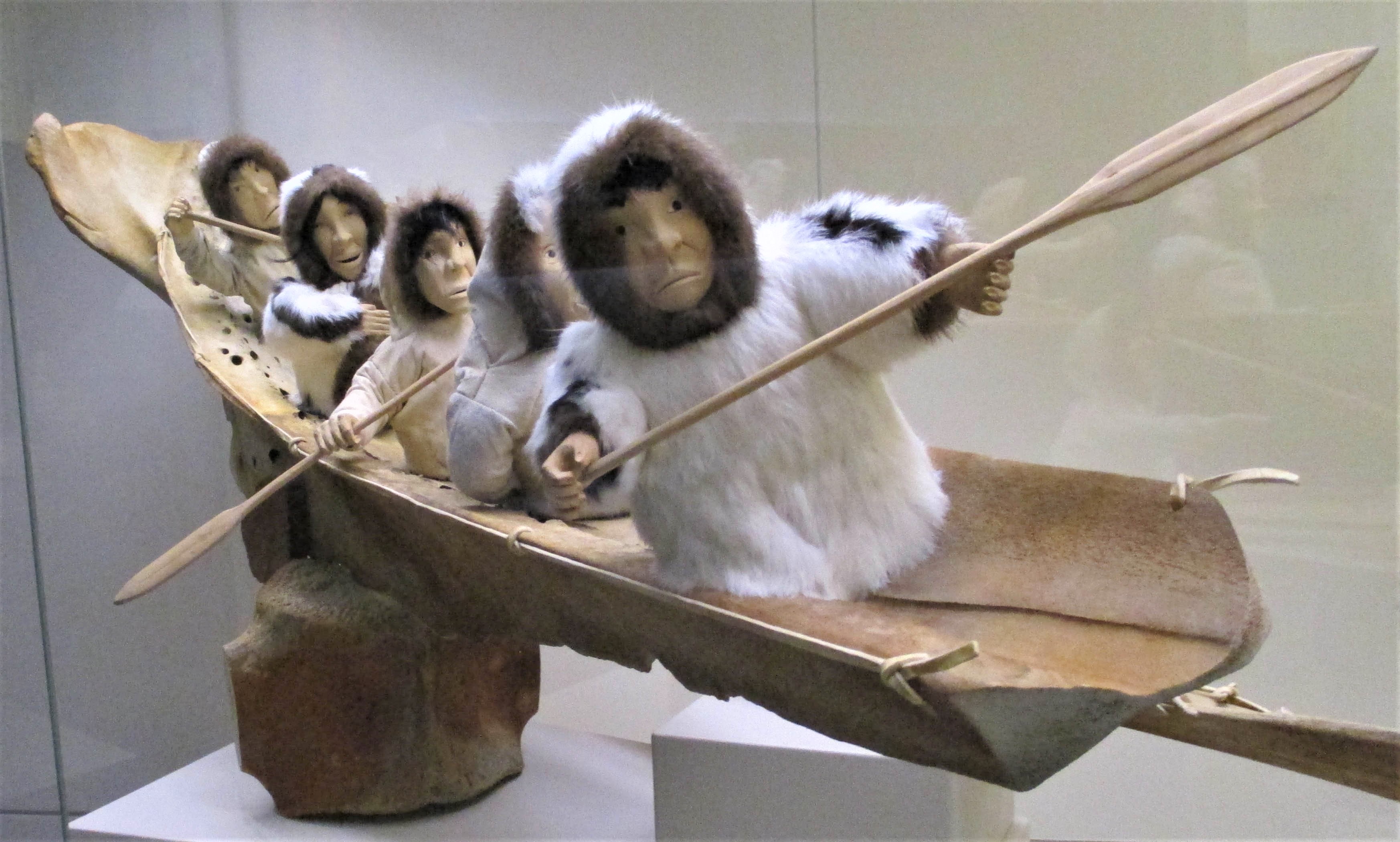 Large whale bone assemblage with rib acting as an umiak with a group of 6 human figures inside, one of whom is a baby in the arms of an adult. Three figures have wooden paddles. Figures wear parkas of caribou and rabbit fur. Faces carved from wood.