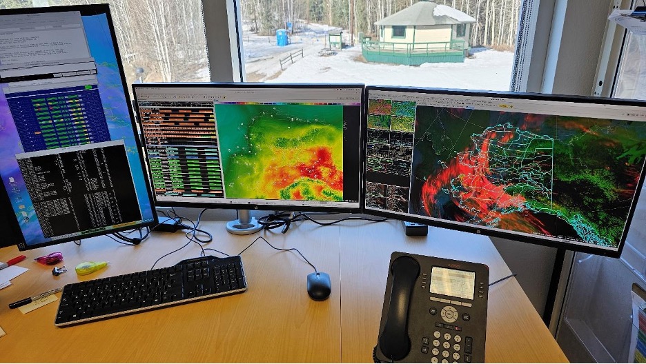 Two computer screens on a desk showing Alaska weather maps, with a ski hut visible outside the window