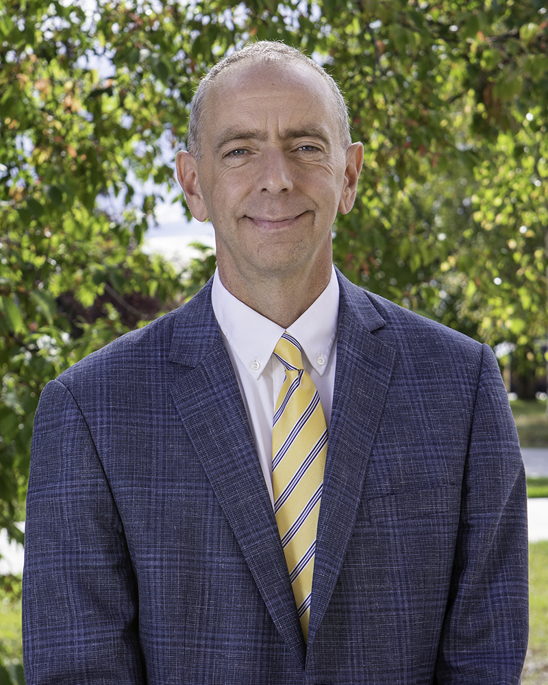 A man stands outside on a spring day, wearing a blue blazer over a white shirt with a yellow, blue, and white striped tie.