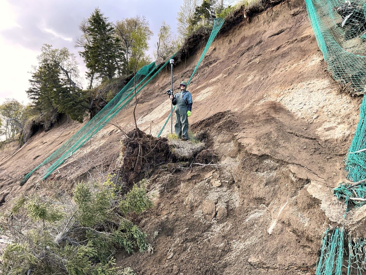 A man holding a pole with a device on top stands on a patch of grass halfway up an eroding hillside. Plastic netting hangs from the top of the hill in several spots.