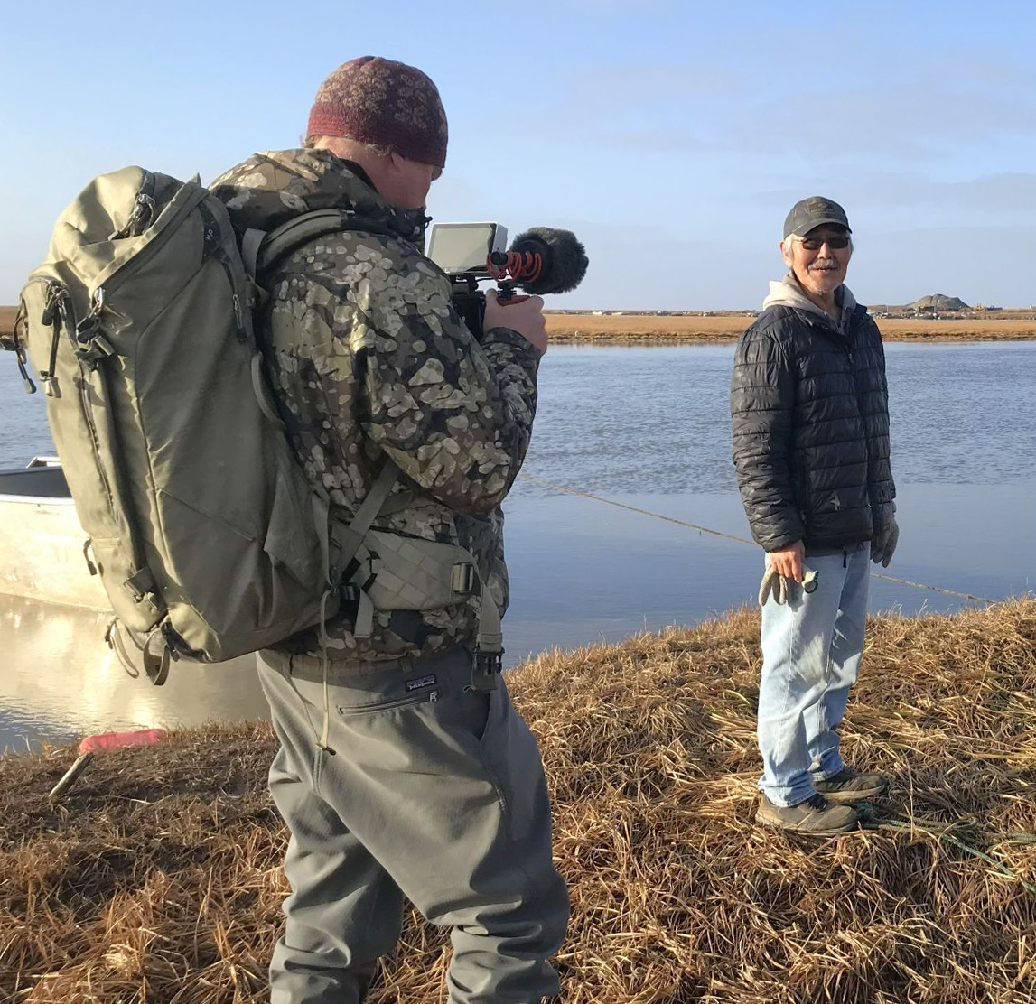A man wearing a backpack and holding a camera and microphone films a man standing on a riverbank.