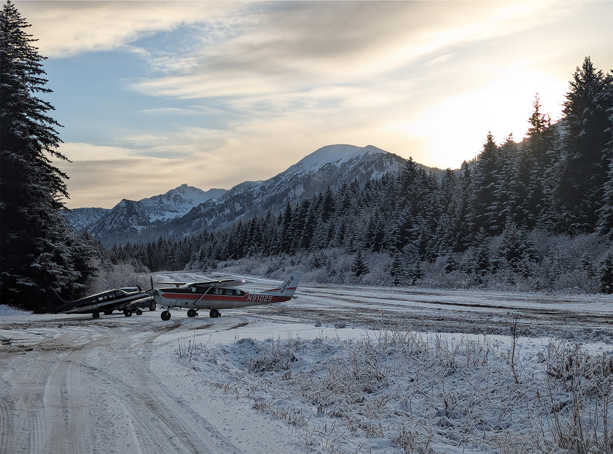 Two small Cessna airplanes are parked beside a rural Alaska airstrip with spruce trees and mountains in the background. Light snow covers the ground, trees, and mountains. The mountains are backlit by a twilight winter sun.