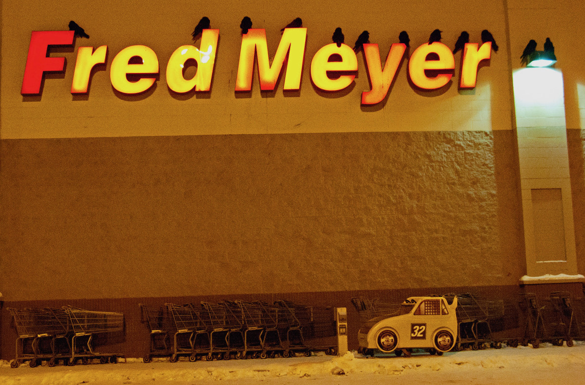 Ravens sit atop signage letters on the exterior wall of grocery story.  Shopping carts on a snowy walkway sit below.