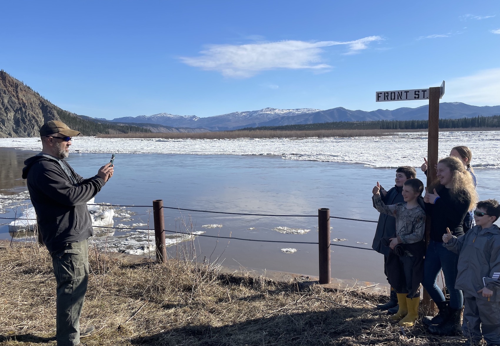 A man, at left, uses a cell phone to take a photo of a group of children gathered at right. Behind them, a river runs partially full of ice.