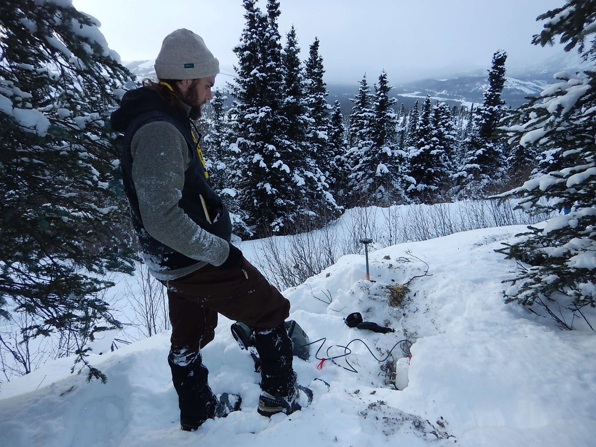 A person stands on a snowy forested hilltop looking down at a piece of equipment partially buried in snow.