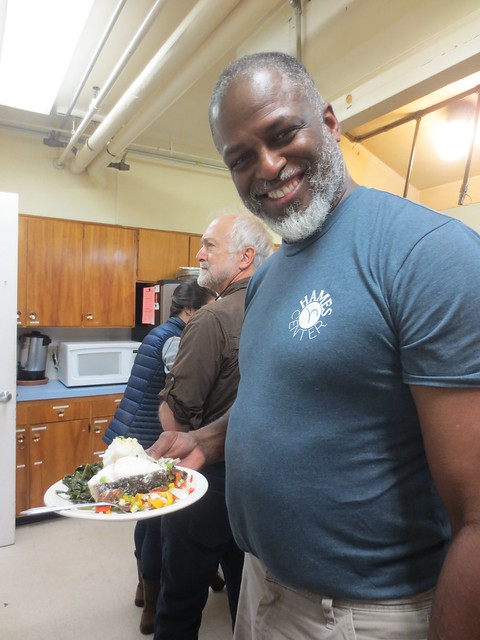 A smiling man stands in a kitchen holding a paper plate filled with food.