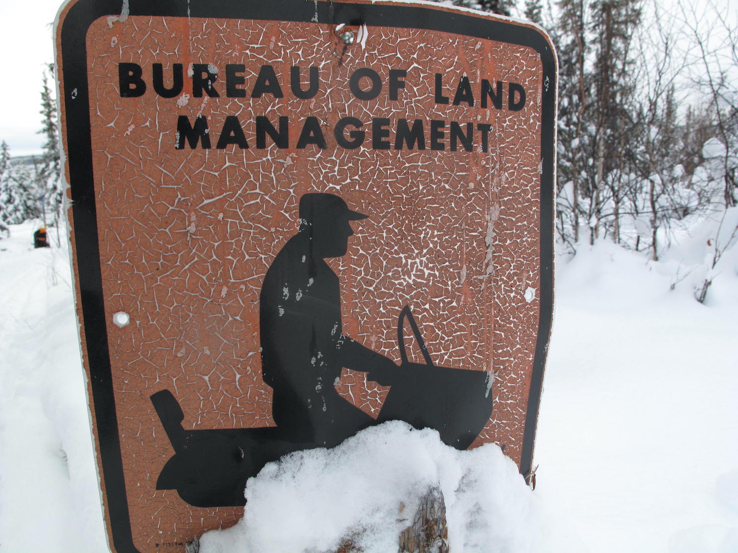 A brown winter trail sign features a black silhouette image of a person riding an old-style snowmachine.