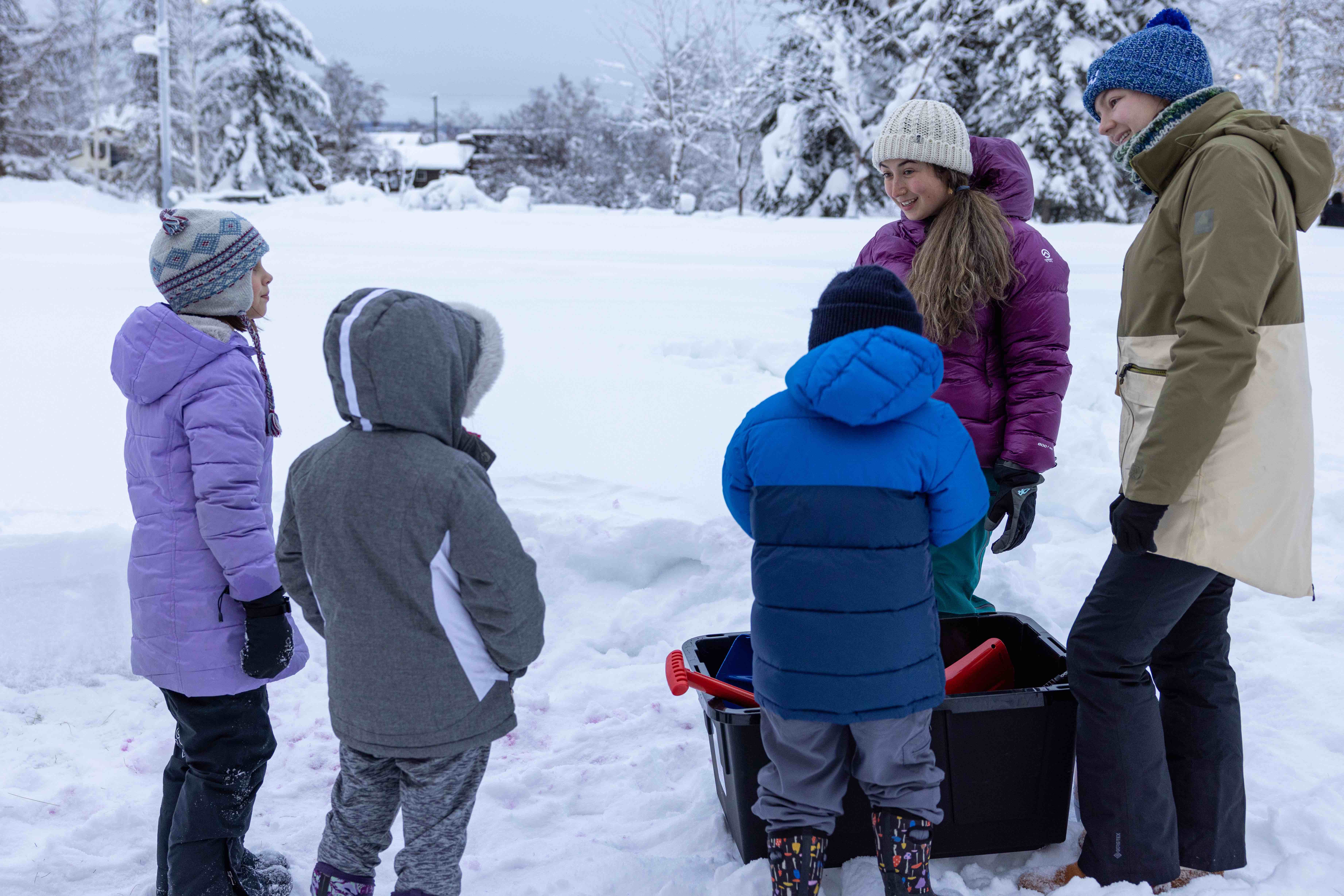 Three elementary-aged children and two adult women stand in a snowy landscape with a box of shovels and other tools.