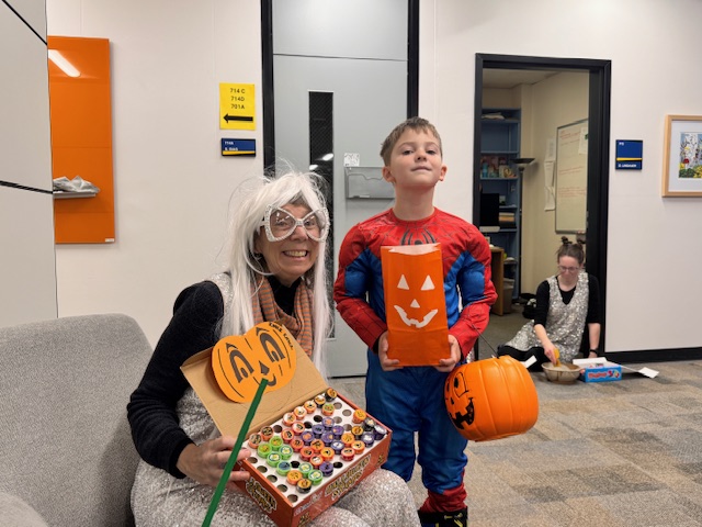 A woman in a white wig with oversized glasses holds a box of candies decorated with a jack-o'-lantern. A boy in a Spider Man costume holds a trick-or-treat bag.