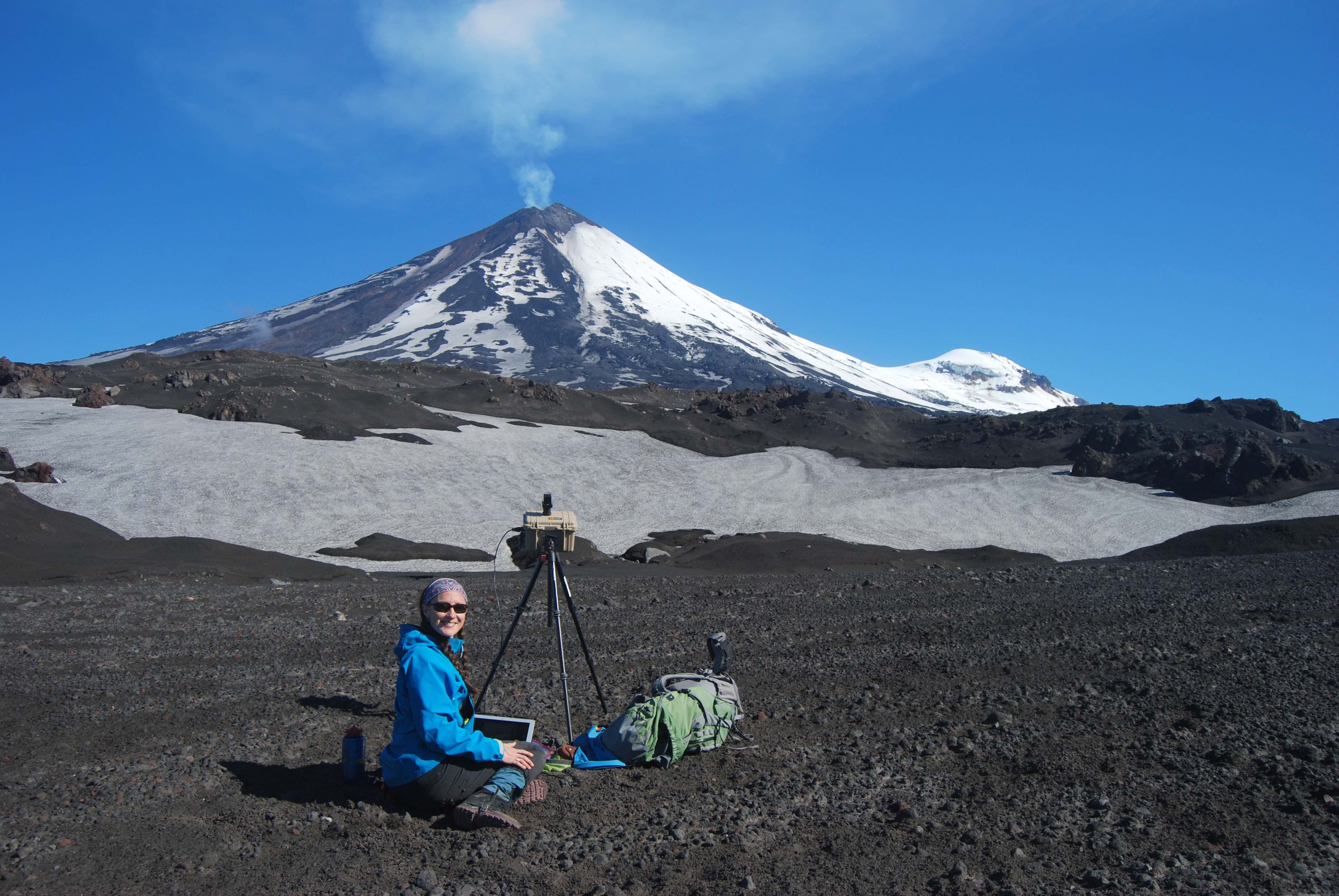 A woman wearing a blue jacket sits on the ground next to a tripod with a volcano in the background. 