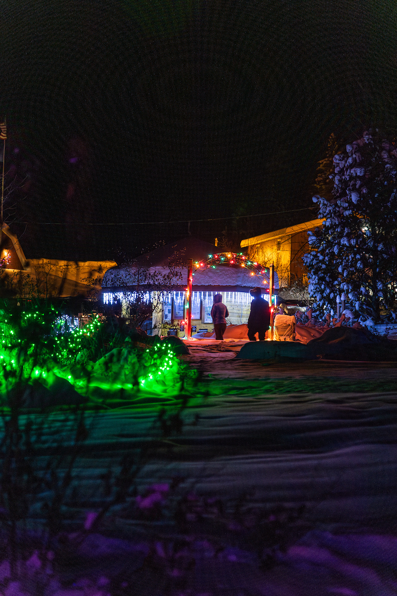 Two people walk through a nighttime landscape with snow and buildings illuminated by colored lights.