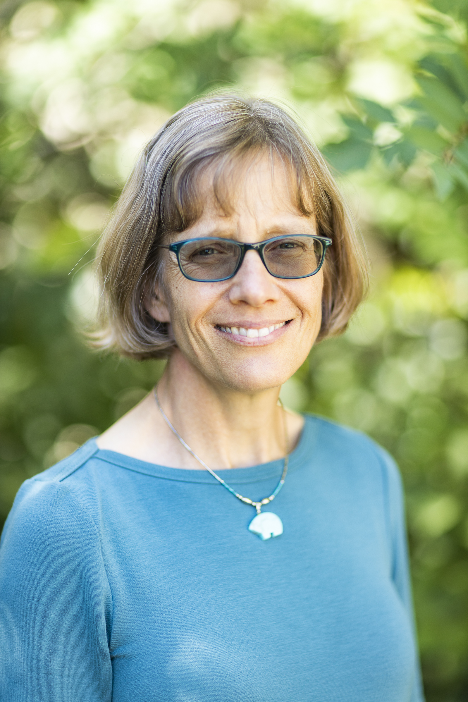 A woman wearing a blue shirt, polar bear pendant and glasses outside during summer on the UAF campus.