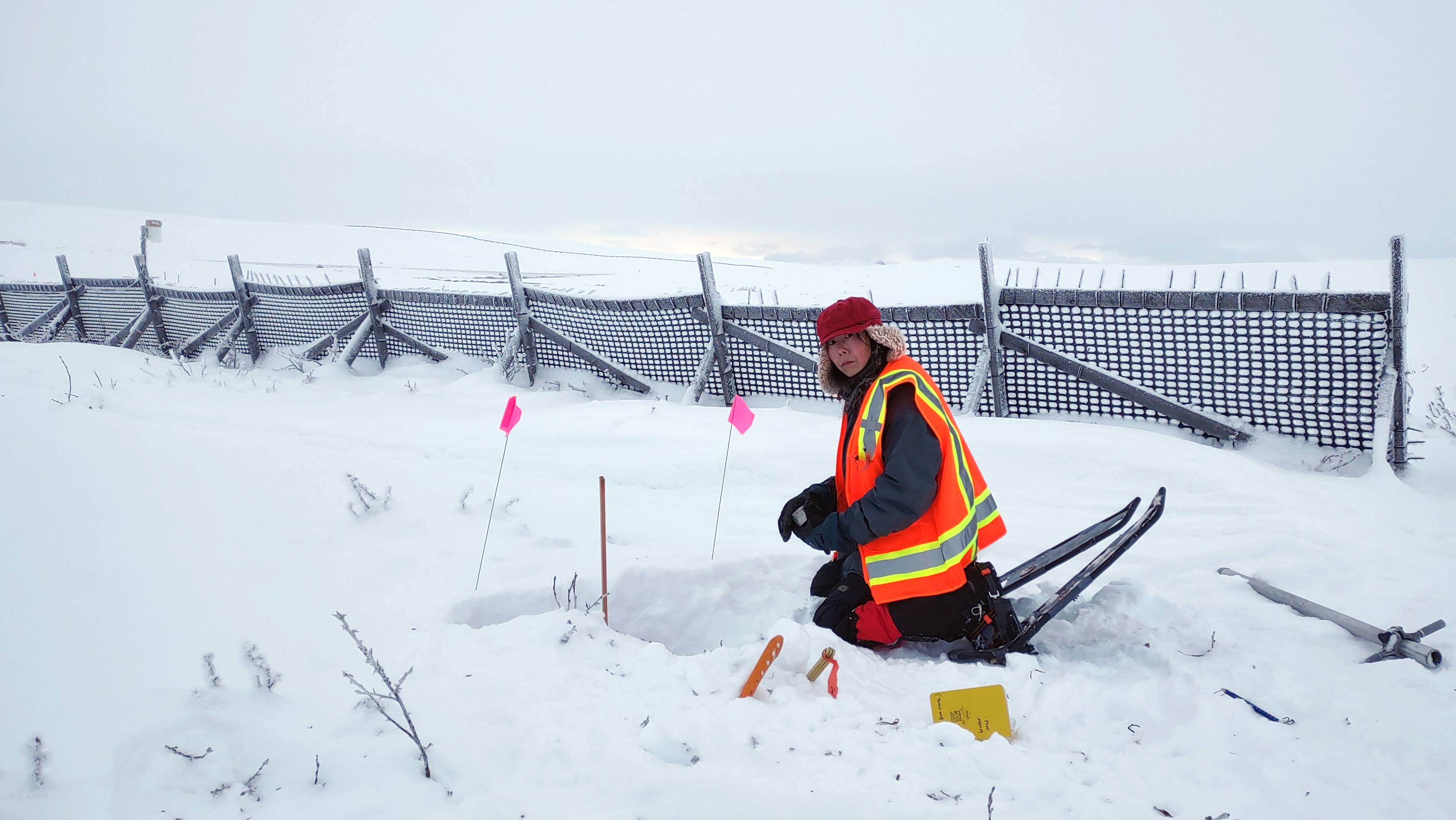 a person in an orange vest crouches in the snow with tools nearby.