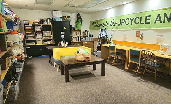 A workspace with a long work counter, sewing machine, yellow sofa and shelves filled with crafting supplies beneath a green 