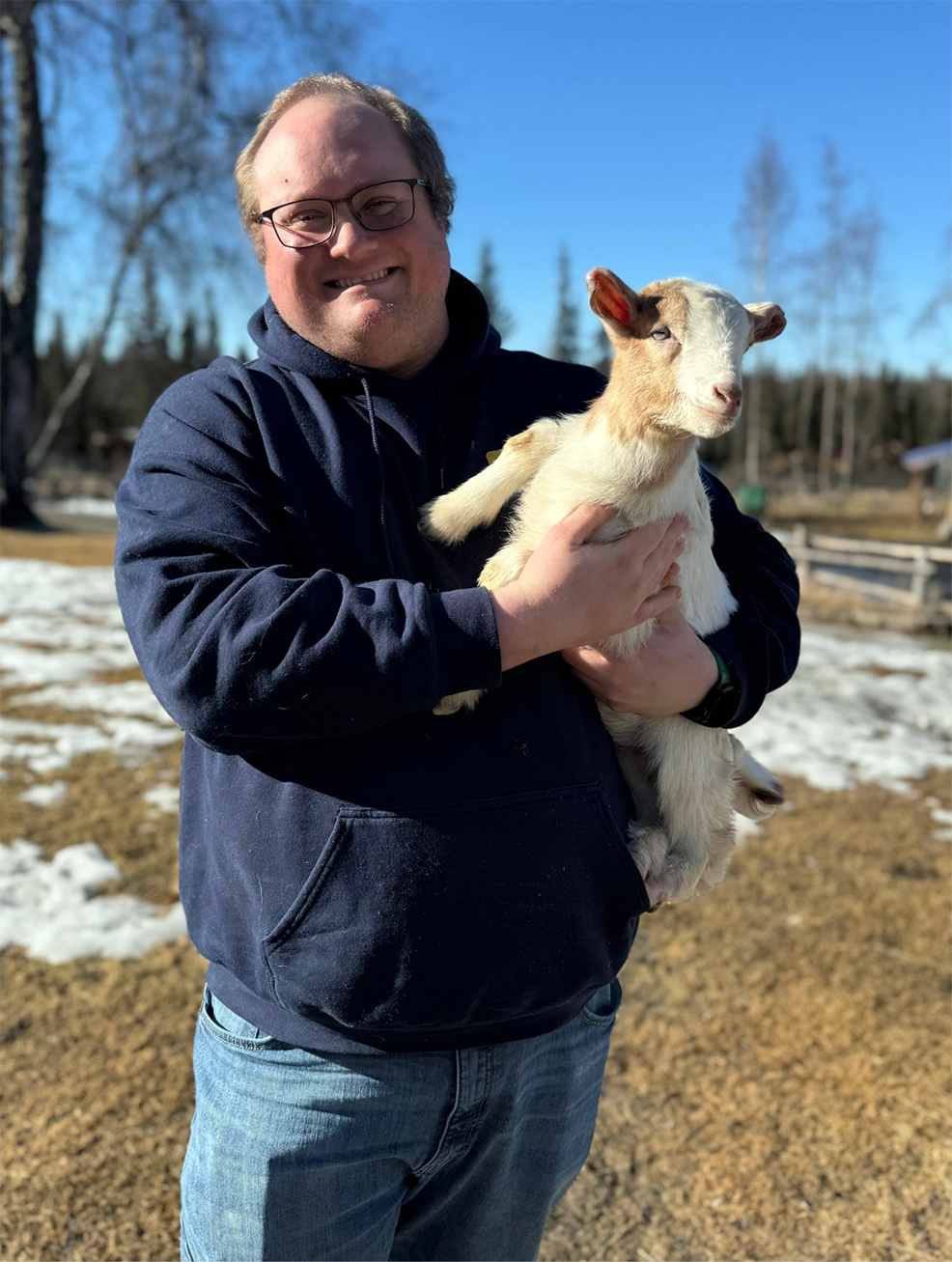 A man, Jim Vinyard, holds a baby goat while standing in a field with patches of snow visible.