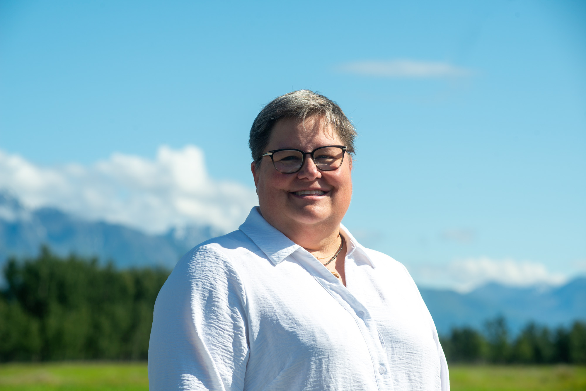 A smiling woman, Jodie Anderson, stands outside under blue skies in front of a snow-covered mountain range.