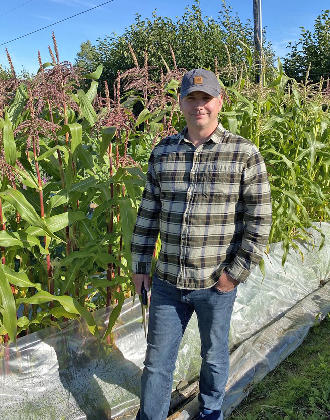 A man in a checked shirt and baseball cap stands next to a row of corn planted through plastic sheeting