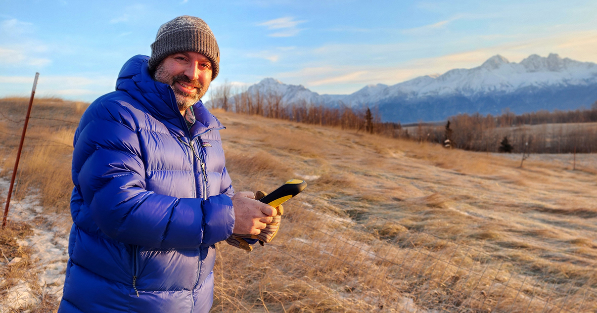 A man in a knit cap and blue puffer jacket holds a GPS reader while standing outdoors near a mountain range