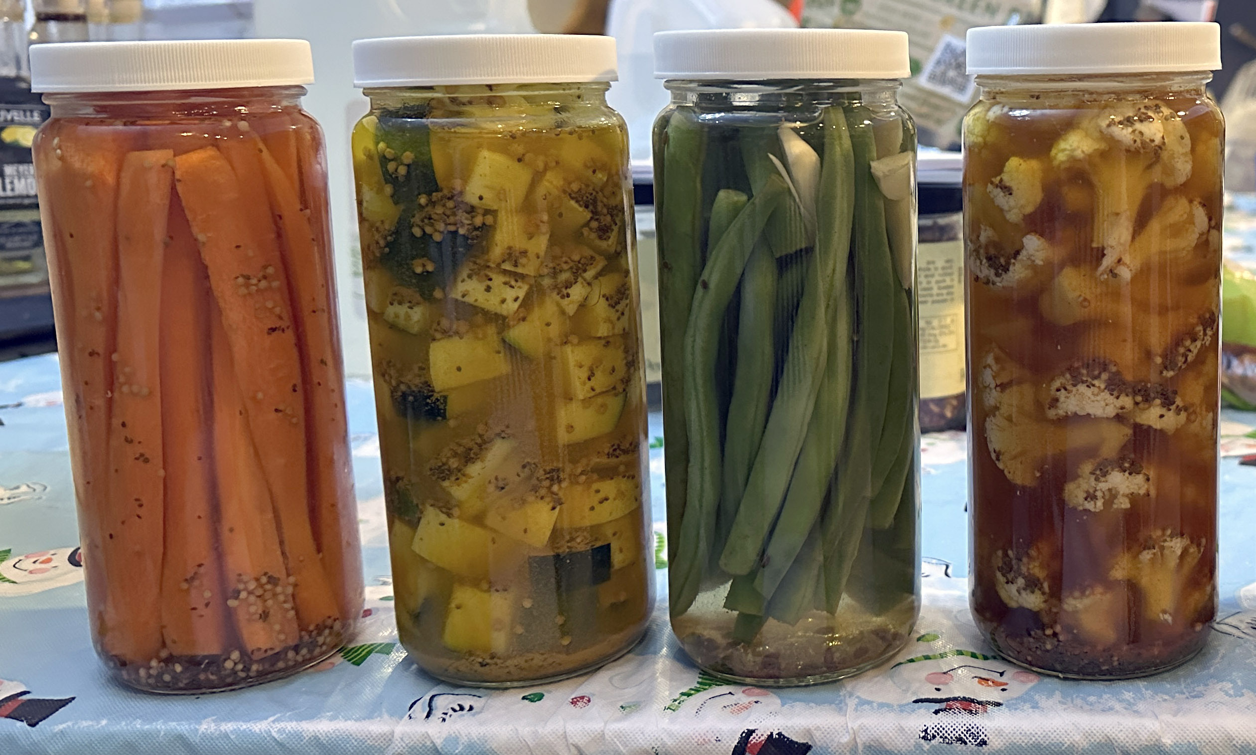 Jars of home-pickled carrots, zucchini, green beans and cauliflower are lined up on a kitchen counter. 