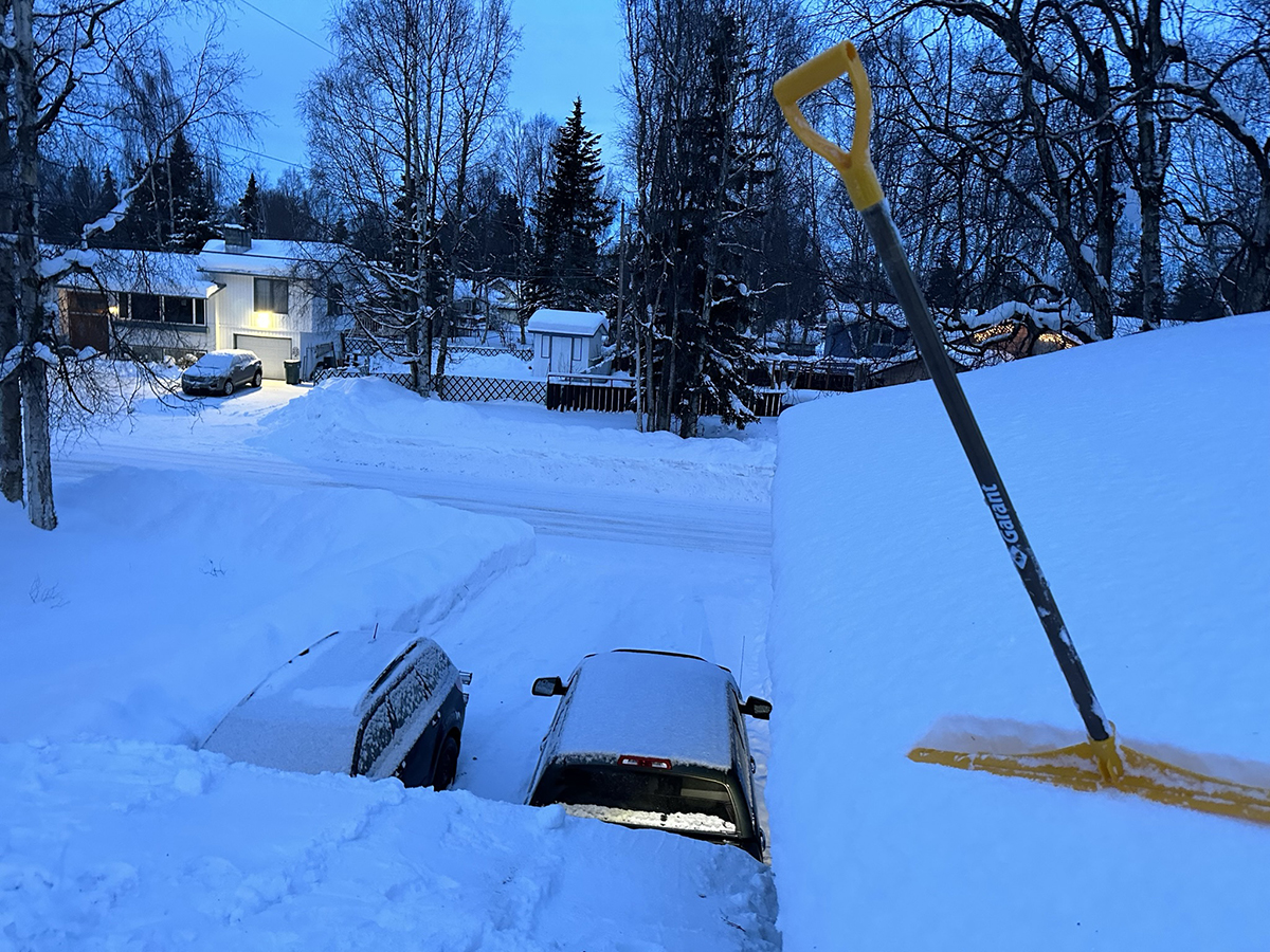 A shovel stands on a snowy rooftop with snow-covered cars and houses in the background.