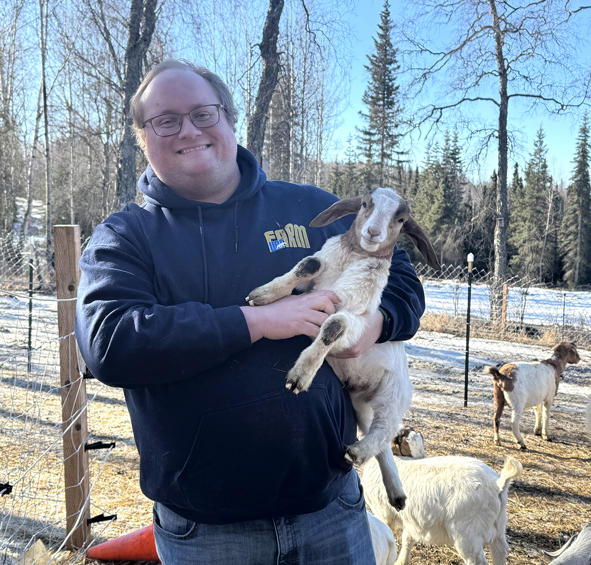 A man holds a baby goat in a wooded field with several other goats in the background.