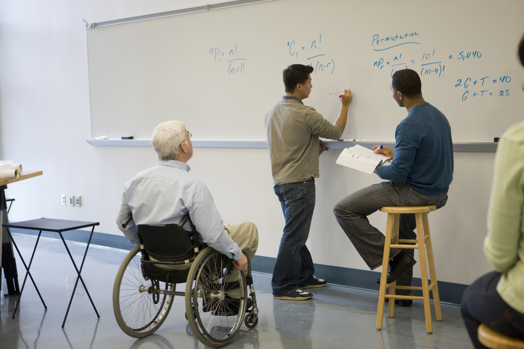 a professor in a wheelchair looks on as two students work out an equation on a whiteboard