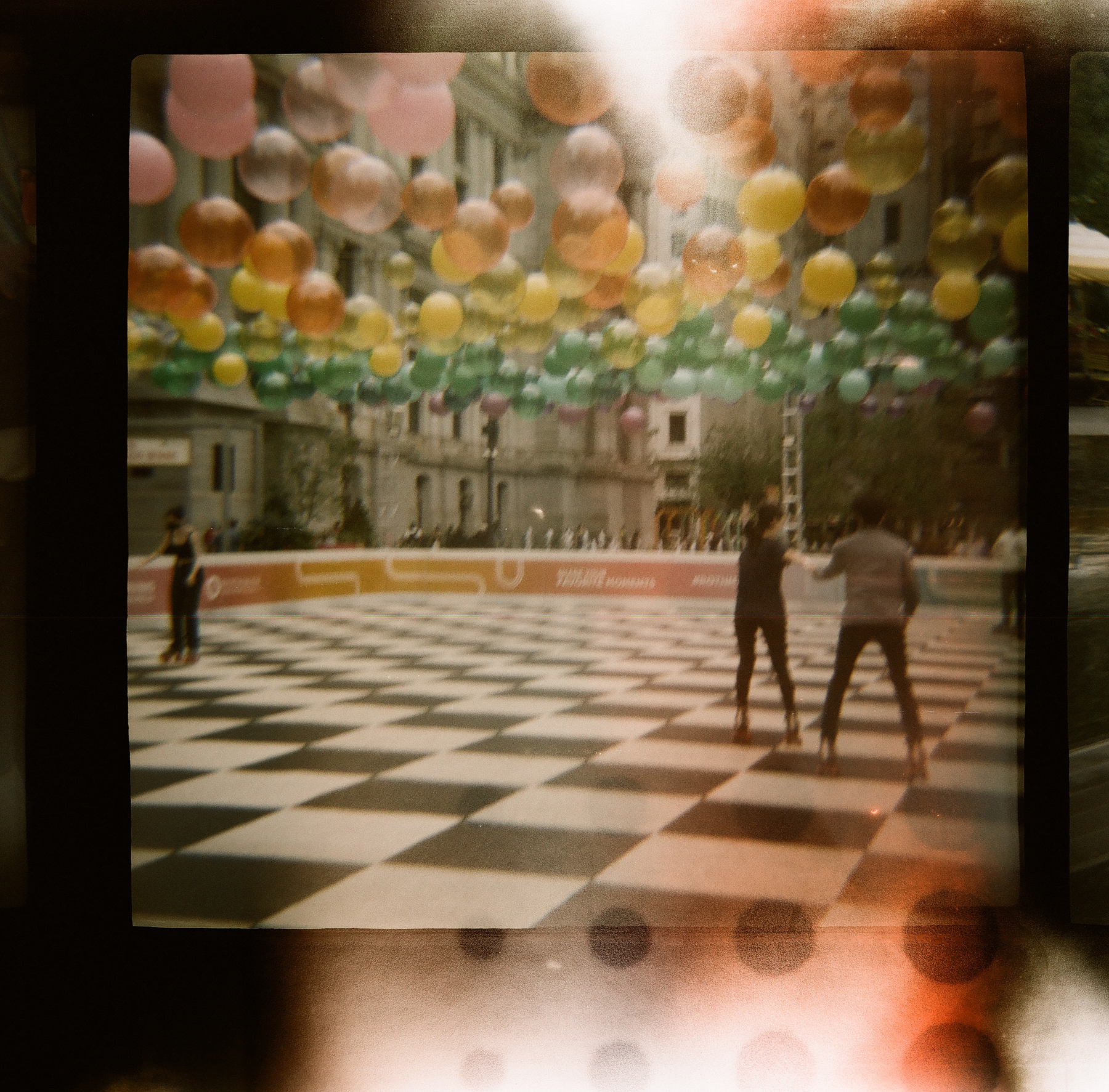 Vintage photograph with light leaks of rollerskaters on a checkerboard rink under a canopy of balloons. Image courtesy of Marina Outwater