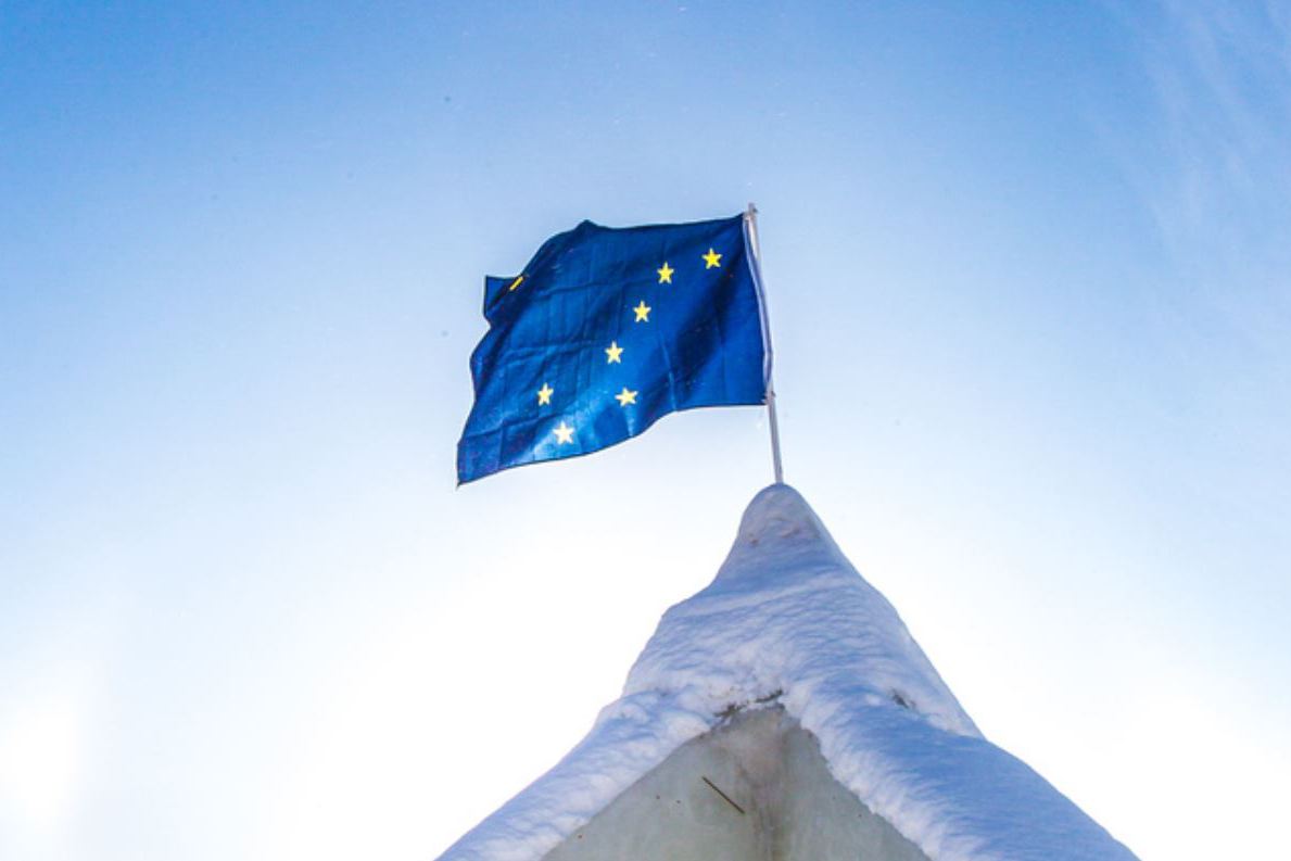 The flag of Alaska waves on top of an ice arch in front of a bright blue sky.