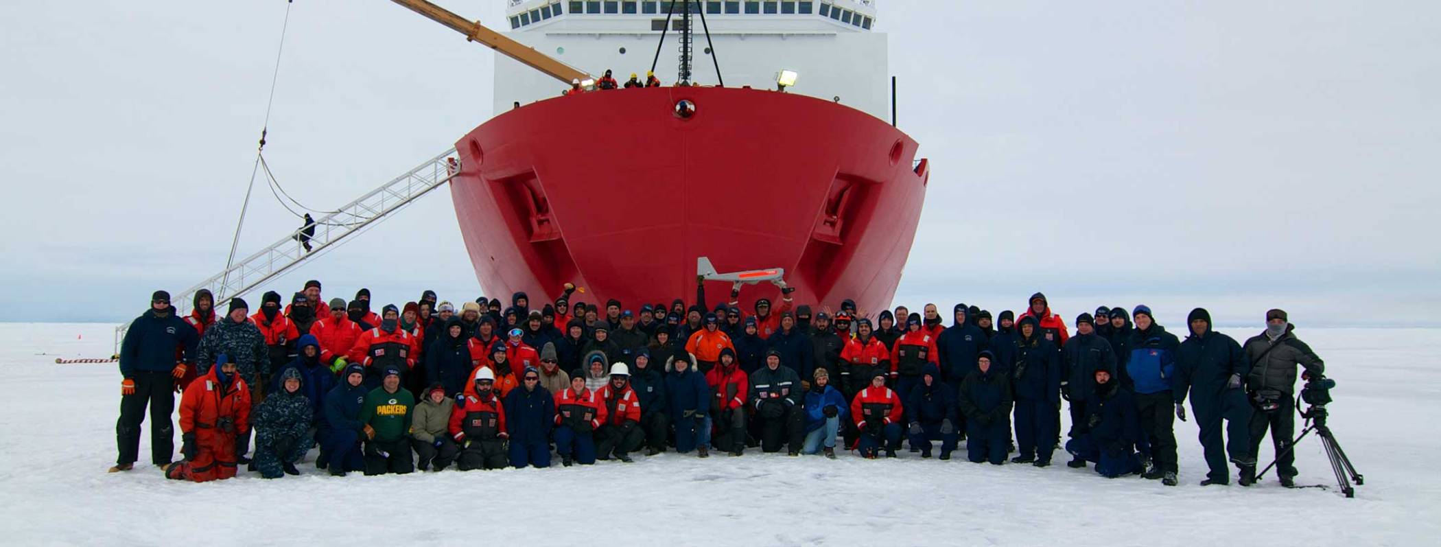 A group photo of people in winter gear standing on the ice in front the bow 1of a research vessel