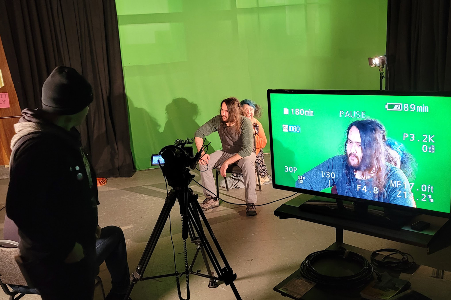 A man sitting in front of a green screen. His image appears on a monitor. UAF Photo by the Department of Theatre & Film