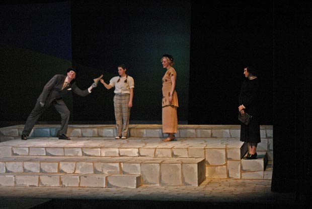 Three women and one man on stage. A man is handing a piece of paper to one of the women as the other two women watch.