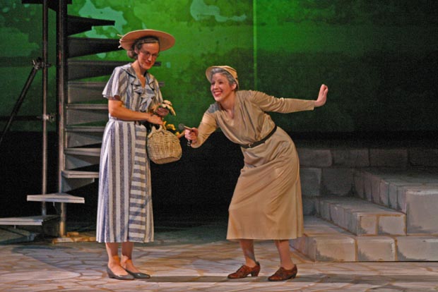 Elderly woman pointing cane towards audience as the woman with a basket of flowers watches by her side.