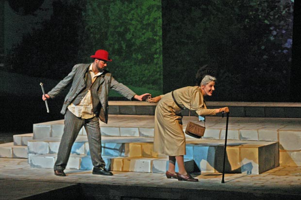On stage an elderly woman using a cane is walking away from a man holding a cane in his hand. The man reaches for the elderly woman.