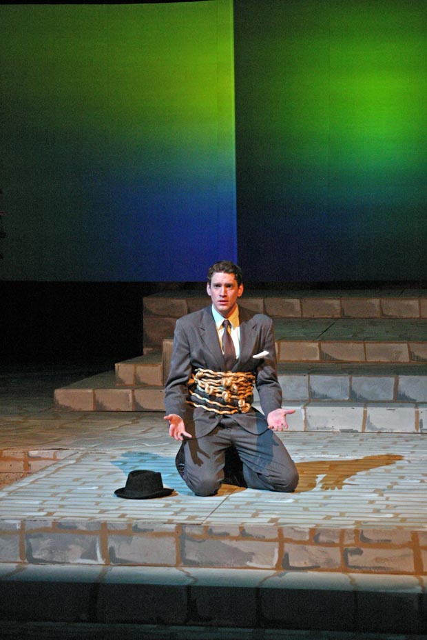 On stage, a man kneels with his waist wrapped in chains and locks, his palms turned upward at waist height.