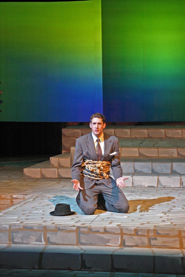 On stage, a man kneels with his waist wrapped in chains and locks, his palms turned upward at waist height.