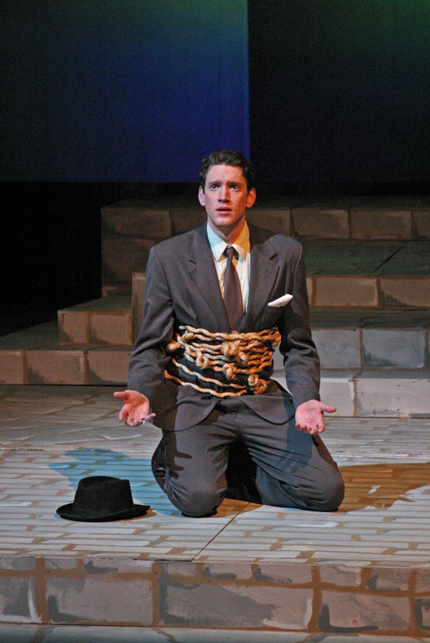 On stage, a man kneels with his waist wrapped in chains and locks, his palms turned upward at waist height.