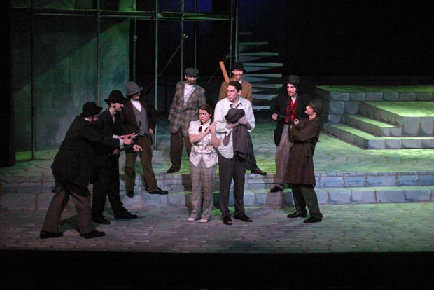 Trapped in the spotlight's glare, a terrified couple confronts a menacing semi-circle of seven men, some brandishing weapons, as uncertainty hangs heavy in the air on stage.