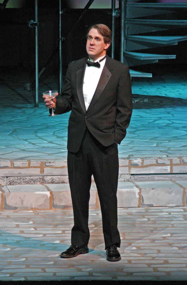 On stage, a man dressed in a tuxedo stands, holding a glass of champagne and looking towards audience with a contemplative expression.