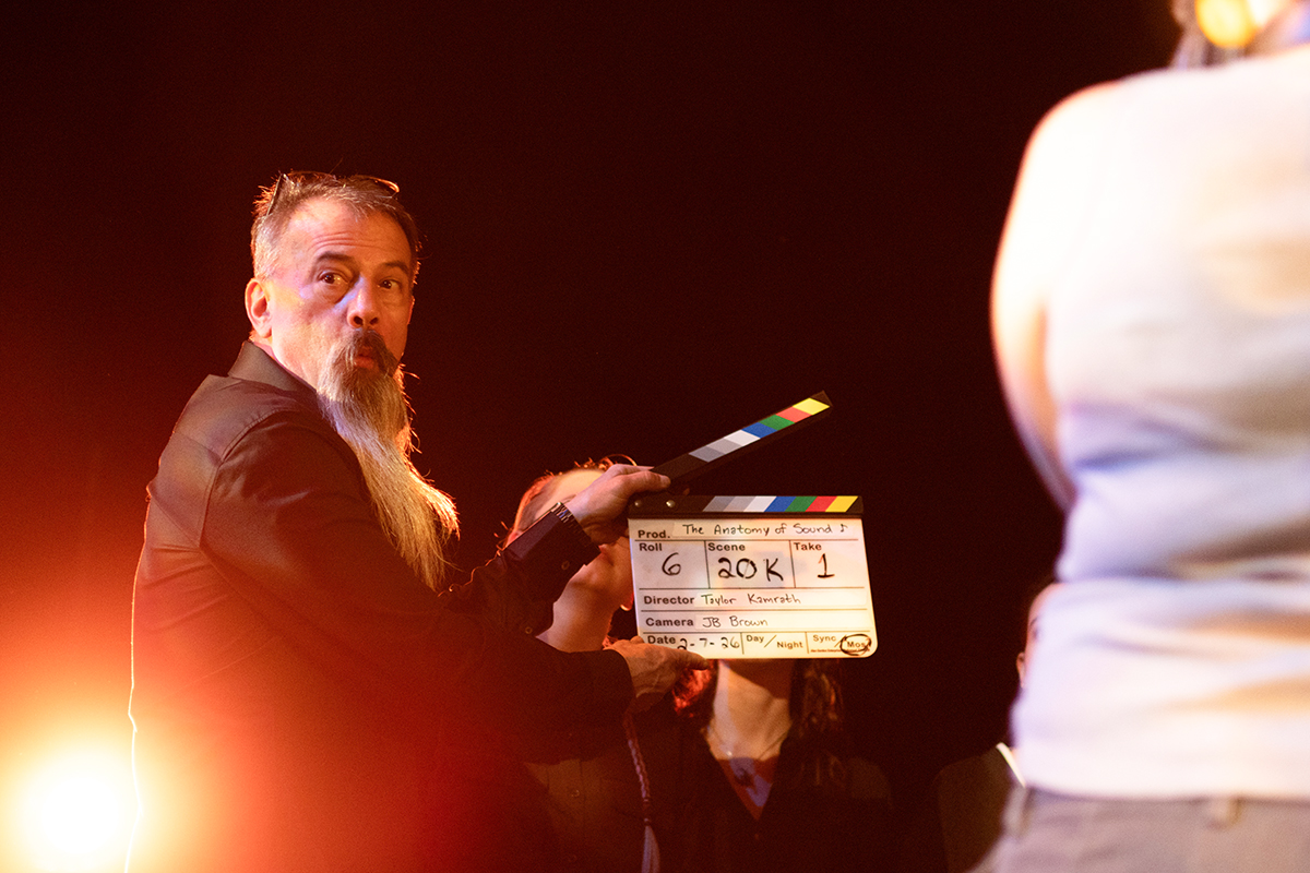 TAOS, Day 2, image 93 Roscoe Archer, backlit by stage lights, holds up a clapperboard to signal the beginning of filming. Photo credit: Eric Marshall