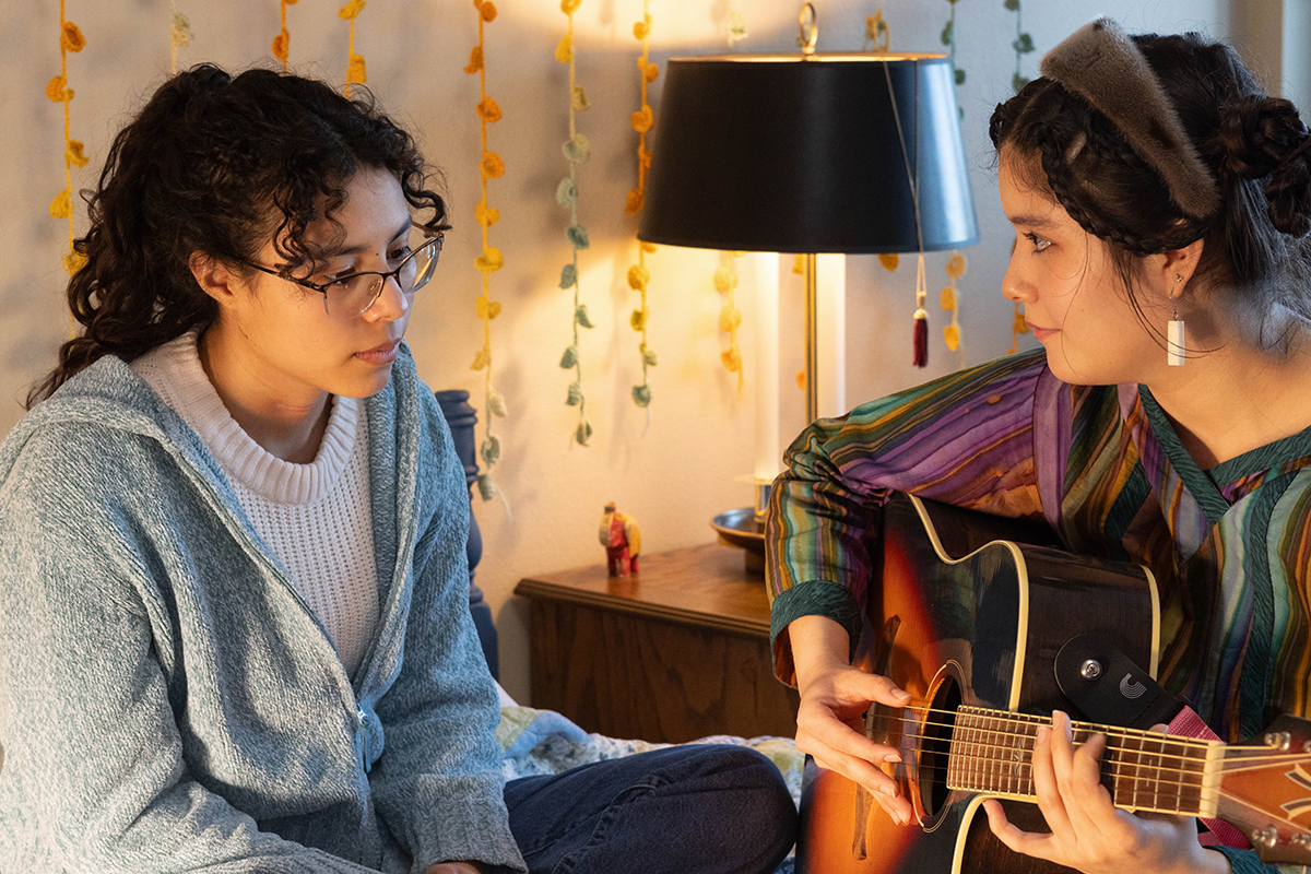 Na’ni’eezh Peter (right, with guitar) sits on a bed alongside Julieth Harvey on the set of The Anatomy of Sound. Photo credit: Eric Marshall