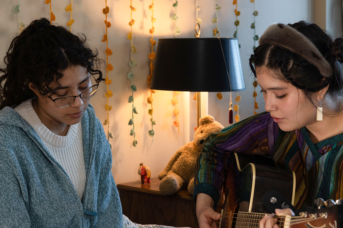 TAOS, Day 8, image 25 Na’ni’eezh Peter (right, with guitar) sits on a bed alongside Julieth Harvey on the set of The Anatomy of Sound. Photo credit: Eric Marshall