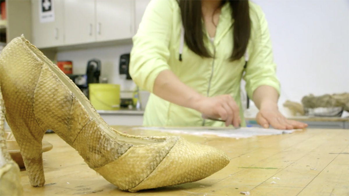To Continue or Be Remembered A high heel made of fish skin sits in the foreground, with the artist at work in the background. Still from To Continue or Be Remembered