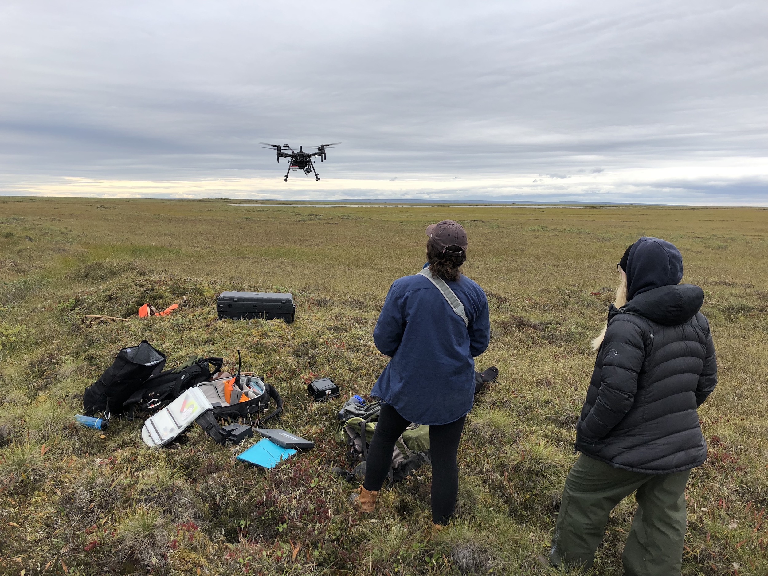 Two women monitor a research-grade drone about 10 feet over the green tundra on a cloudy summer day. The woman on the left controls the drone while the other supervises.