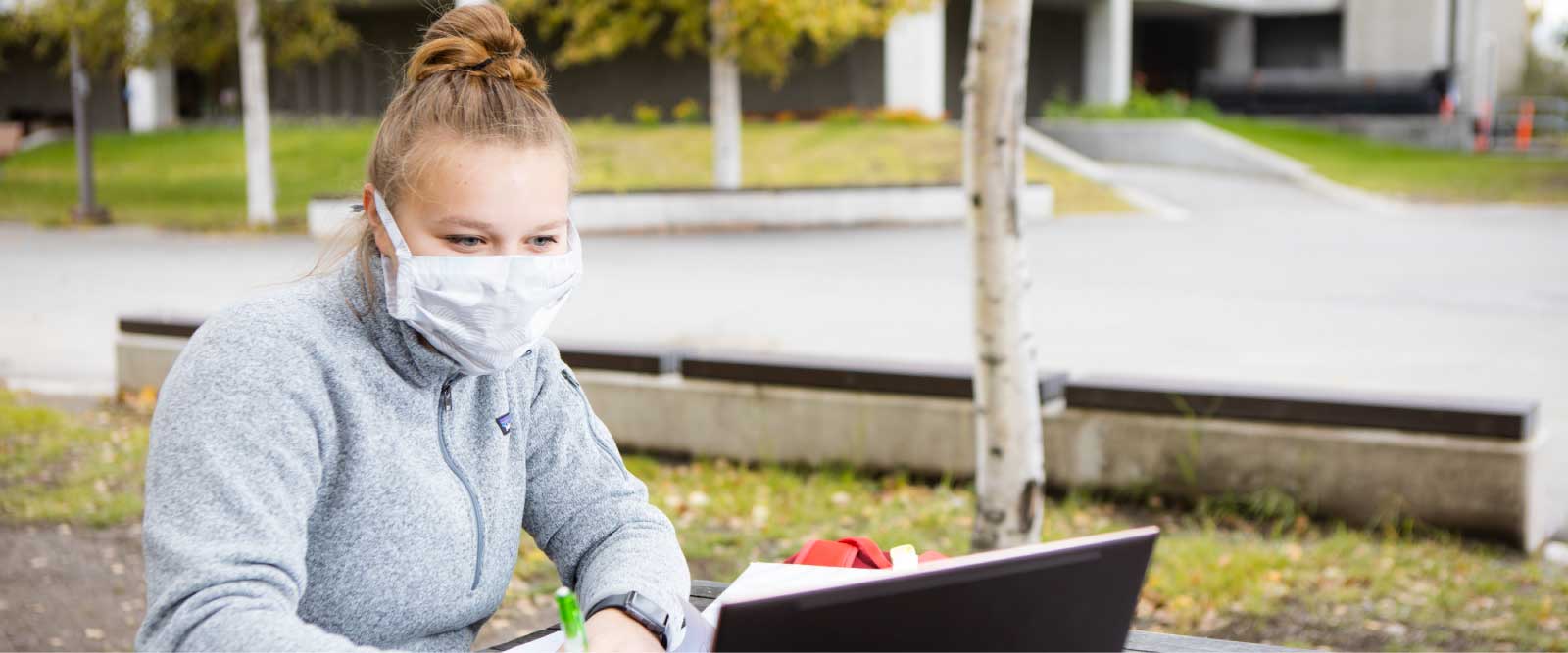 A student wearing a mask sits at a table with a laptop outside the Wood Center