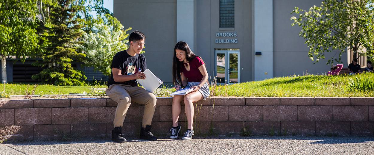 Two UAF student compare notes while seated in Cornerstone Plaza in font of the Brooks Building on the Fairbanks Campus in summer.