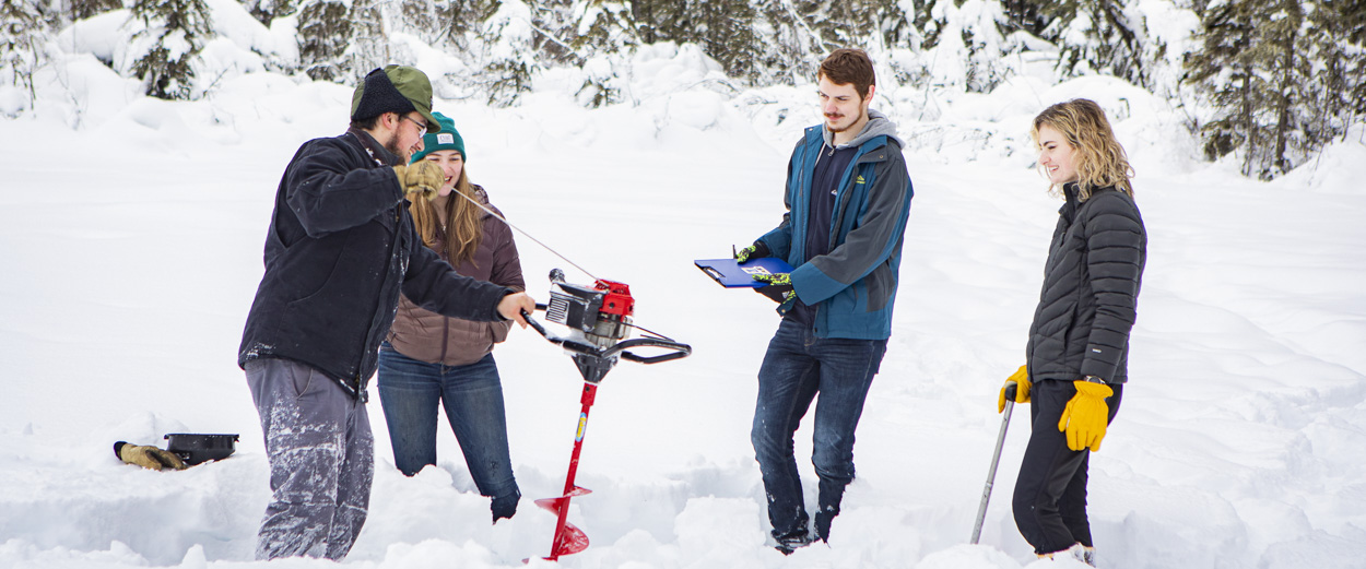 4 UAF students stand on a snow covered lake. One is holding an ice auger, as 3 others look on.
