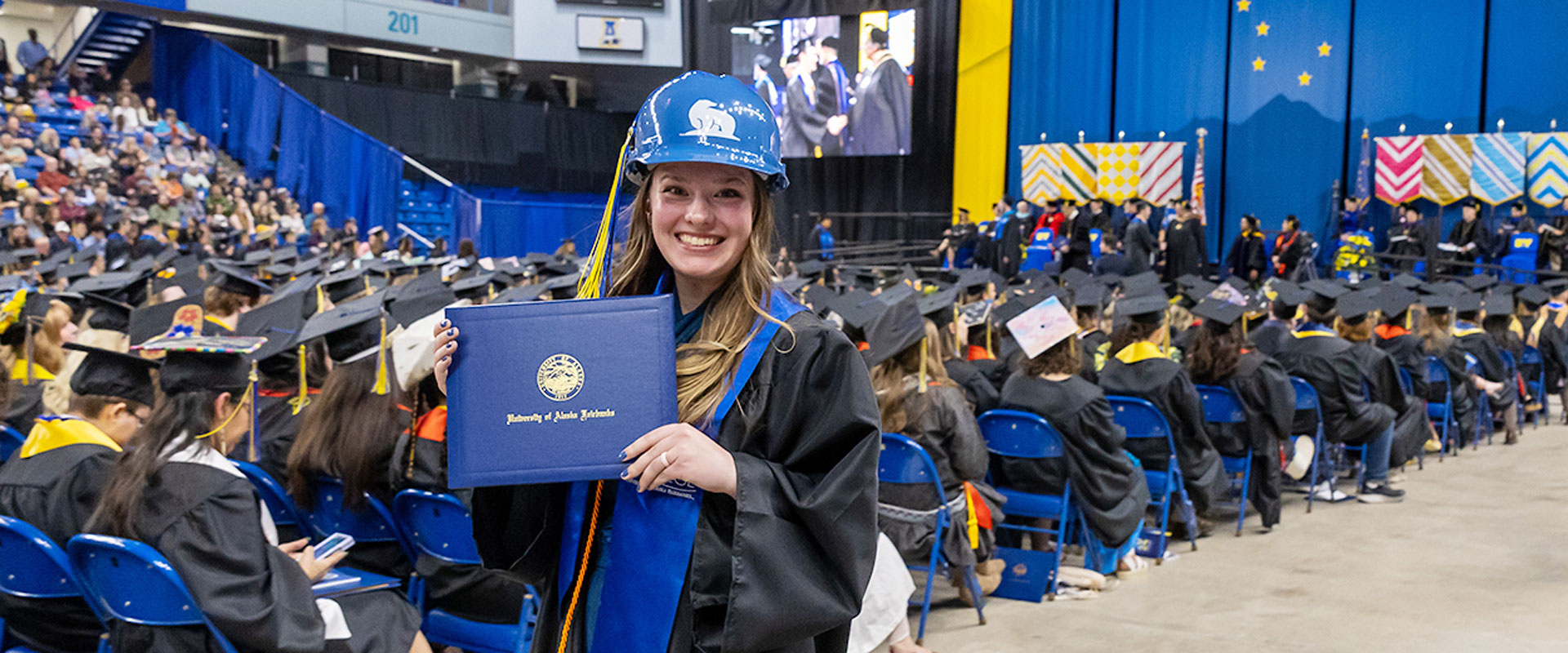 A UAF graduate wearing a hard had holds up a diploma cover at the 2024 UAF Commencement Ceremony.
