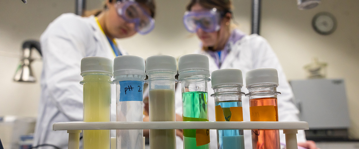 Two students in a lab wearing googles and lab coats, with colorful test tubes in the foreground